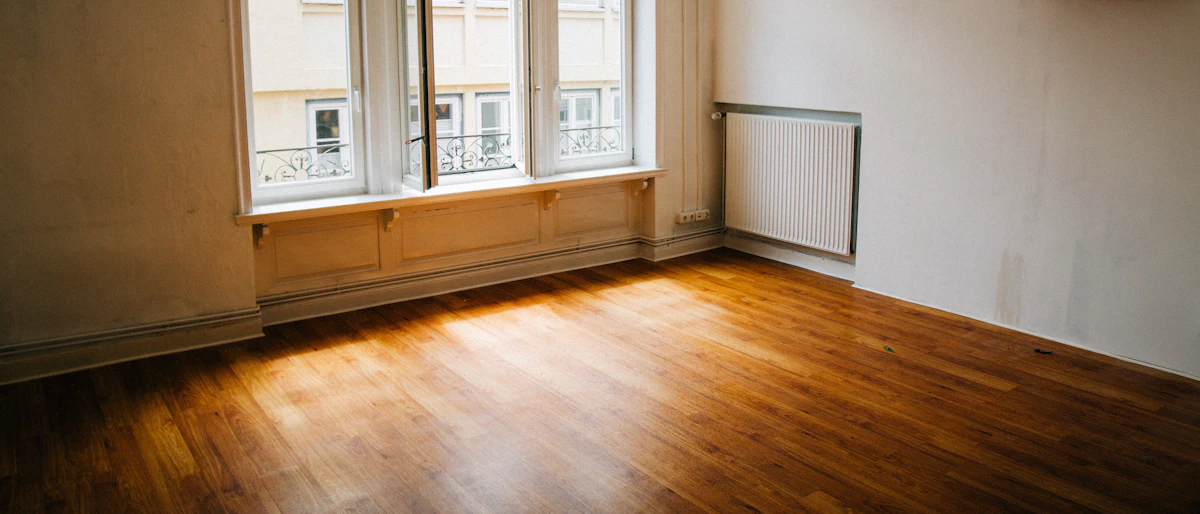 Bright, empty apartment room with wood flooring and a large window, ready for handover